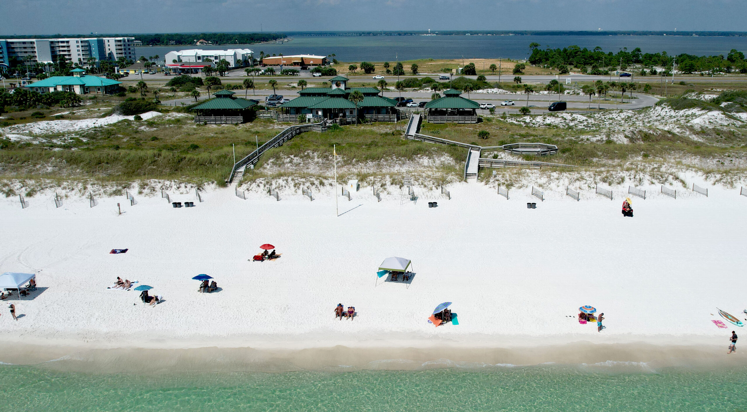 Aerial view of a beach with white sand, a few people under umbrellas near the shoreline, and buildings with green roofs in the background near a bay.