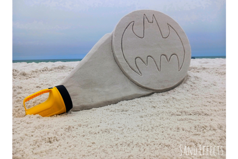 A sand sculpture of a large flashlight with the batman logo illuminated on its front, against a beach backdrop.