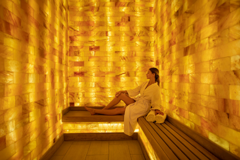 A woman in a white robe meditates in a warmly lit salt cave sauna, sitting cross-legged on a wooden bench.