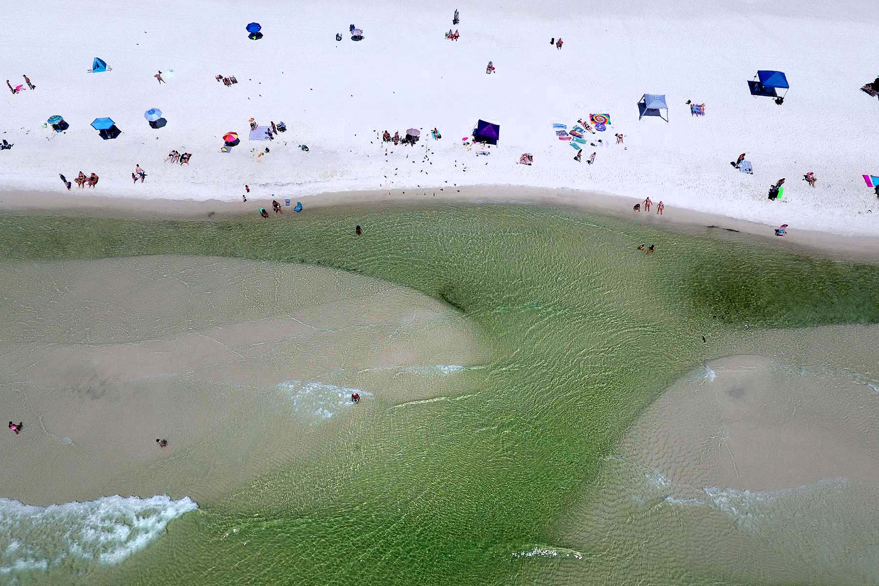 Aerial view of people on a white sandy beach with umbrellas and tents, a green tidal area in the water, and a few swimmers in the shore.