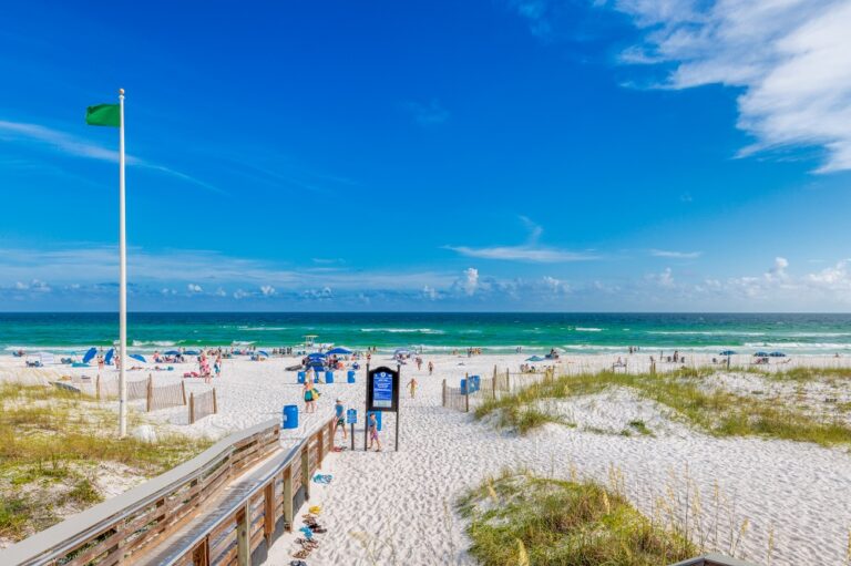 A scenic beach view with white sand, green flag, and several people under umbrellas near the ocean. The sky is clear and blue.