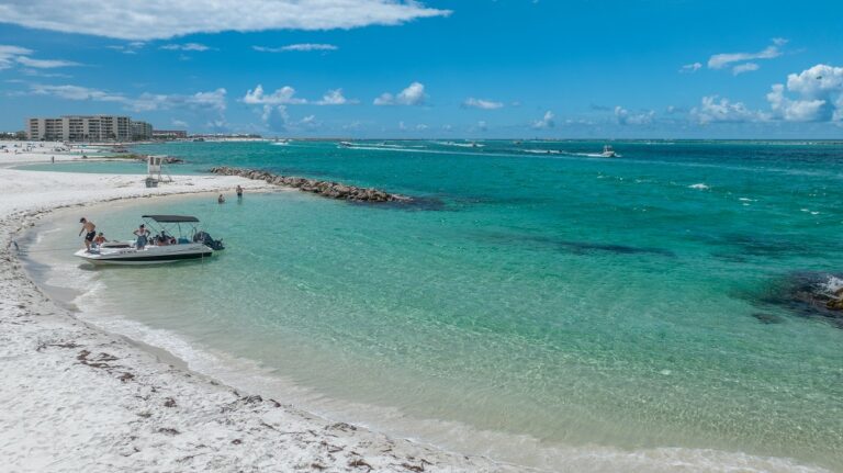 A small motorboat is anchored close to the shore on a sandy beach with turquoise water. People are relaxing on the boat and strolling along the beach under a clear blue sky.