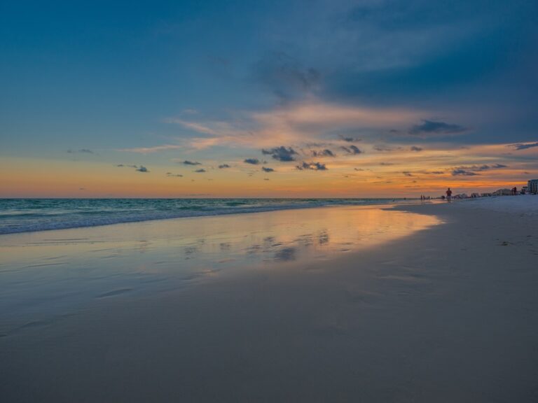 A tranquil beach at sunset with a vast sky reflecting orange and blue hues on the calm water. Scattered clouds enhance the serene atmosphere.