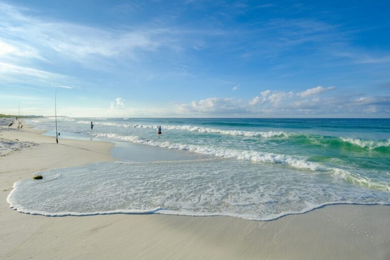 A serene beach scene with gentle waves lapping the shore, a fishing rod in the sand, and people in the distance under a partly cloudy sky.