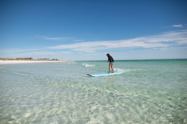 Person paddleboarding on clear, shallow water with a sandy beach and blue sky in the background.