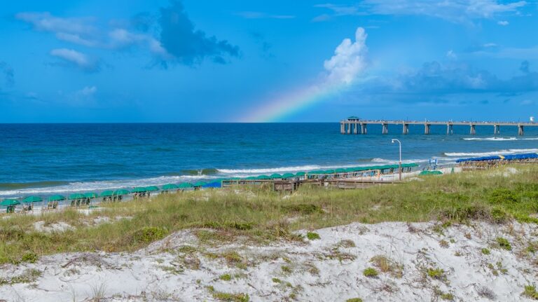 A sandy beach with grassy dunes, a row of umbrellas, a wooden pier extending into the ocean, and a faint rainbow in the sky.