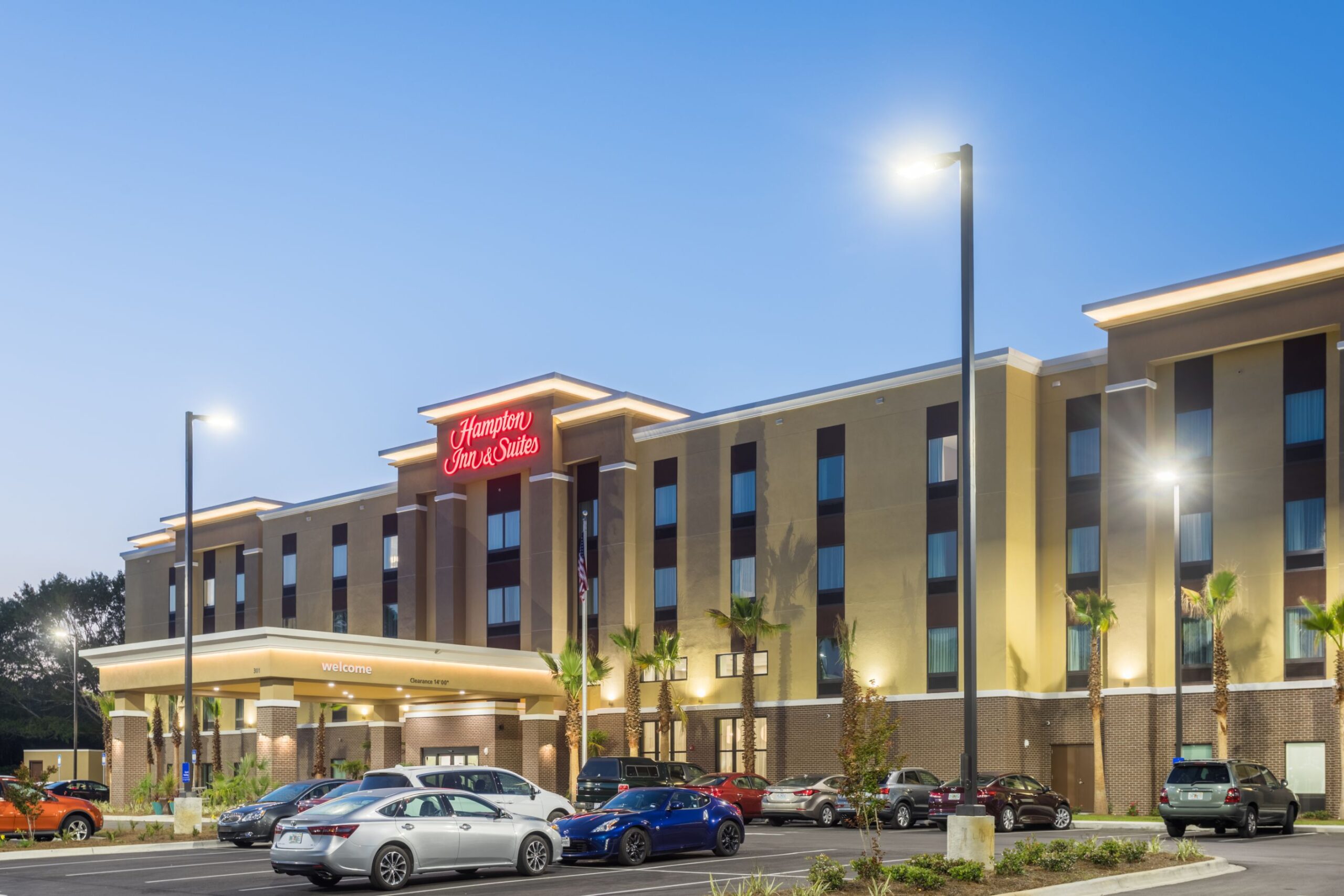 A Hampton Inn & Suites hotel building with illuminated signage at twilight, surrounded by parked cars and palm trees in the parking lot.