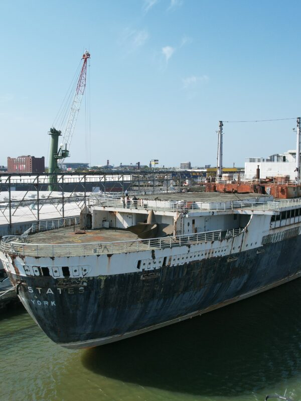 Large, weathered ocean liner docked at an industrial port under a clear blue sky.